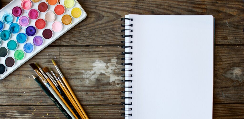 paint brushes next to drawing book and water color palette